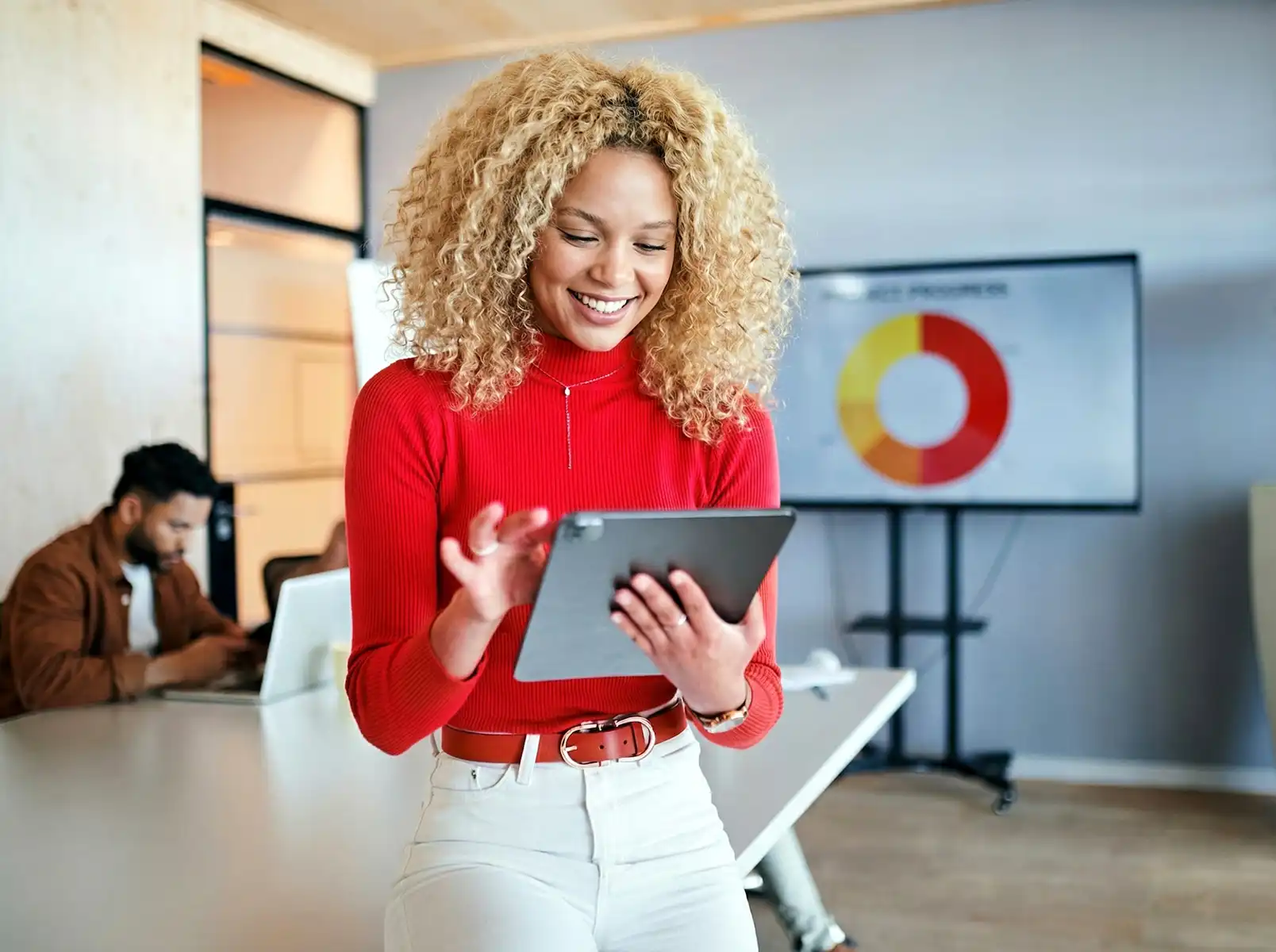 A woman with a tablet in a meeting room, with a man working on a laptop in the background and a large pie chart on the wall.